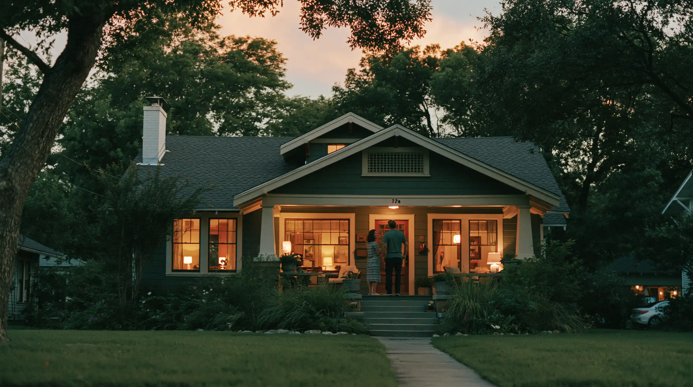 A modern craftsman-style home glowing warmly at dusk, with a couple standing together on the lit front porch.